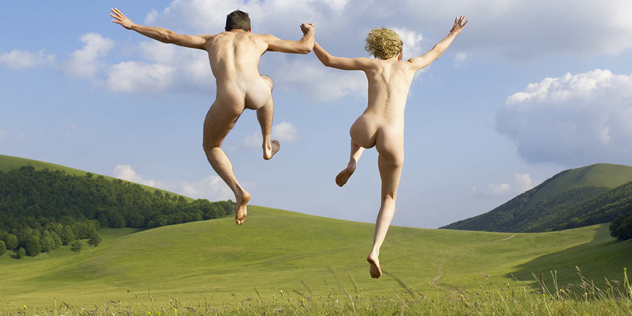 Nudist couple holding hands and leaping into the air as they run across a grassy field