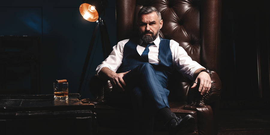 Mature handsome dominant male with a beard and wearing a navy suit reclining in a leather ottoman drinking a spirit