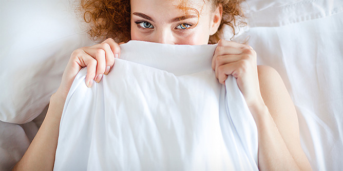 Shy woman with curly red hair holding a sheet up to partially hide her face