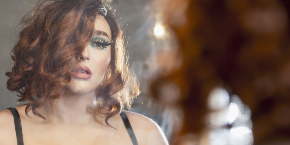 Cross dresser in makeup, wig and little black dress admiring reflection in mirror