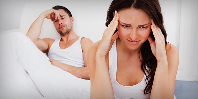 Woman with her head in her hands sitting on edge of bed where her partner is lying