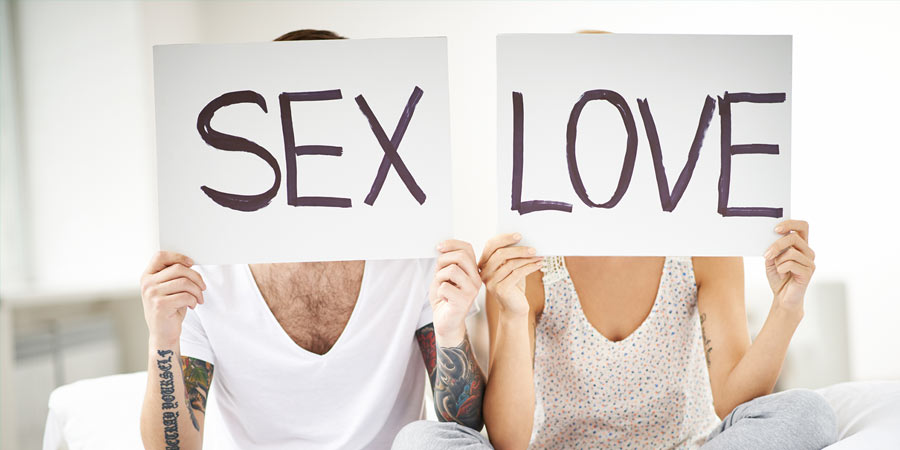 Man and woman sitting cross legged on a bed holding signs with SEX and LOVE displayed