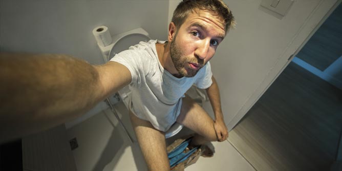 Man sitting on a toilet with his jeans around his ankles while he takes a selfie