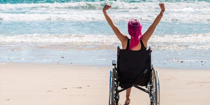 Disabled woman with bright pink hair sitting in a wheelchair on a beach watching the ocean