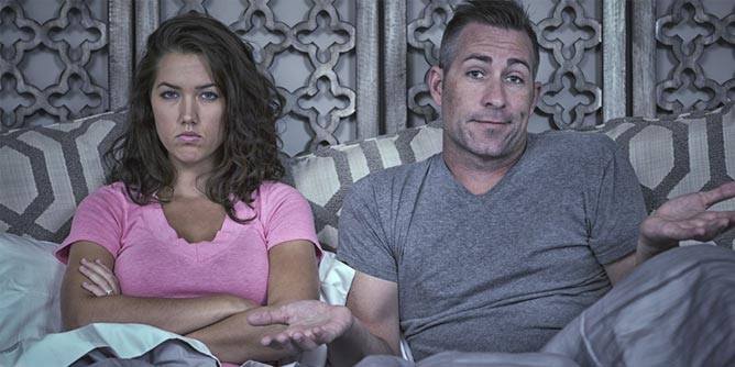 Angry couple sitting in bed with the man shrugging and the woman with her arms crossed