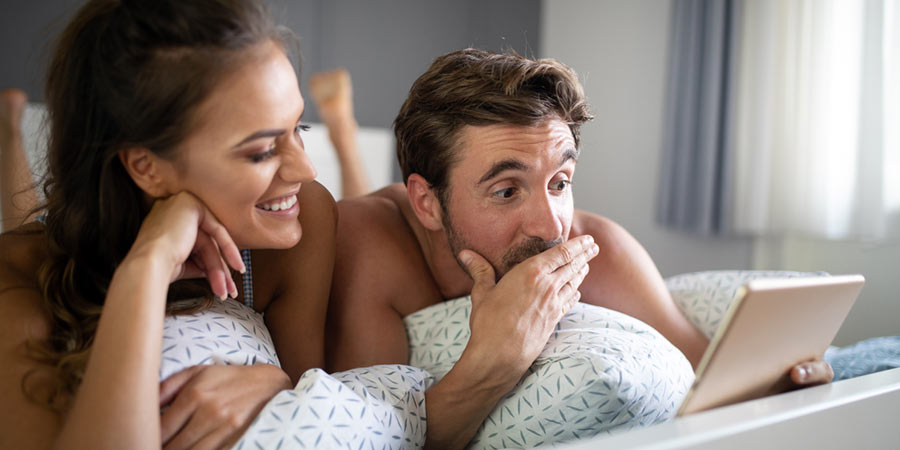 Couple lying in bed watching porn on a tablet with the man looking shocked