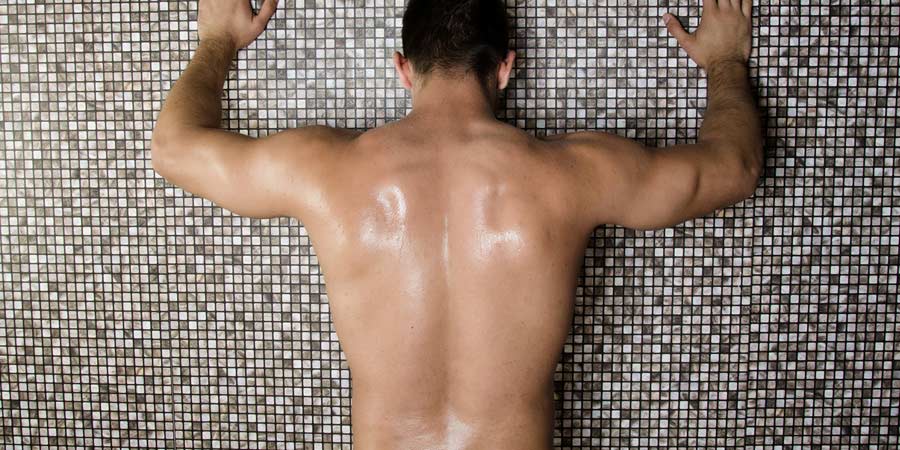 Naked man standing with back to the camera leaning up against a tiled shower