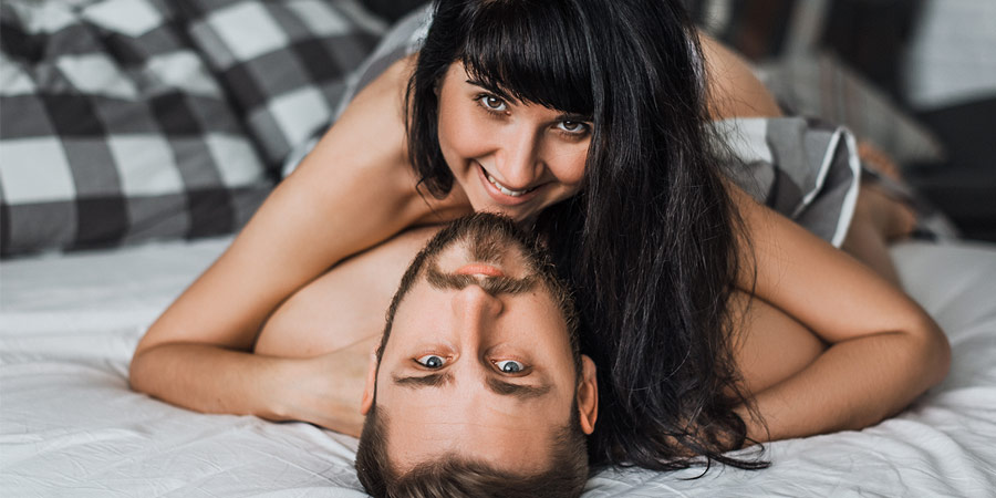 Sexy young woman with long dark hair lying on top of her partner in bed and smiling at the camera