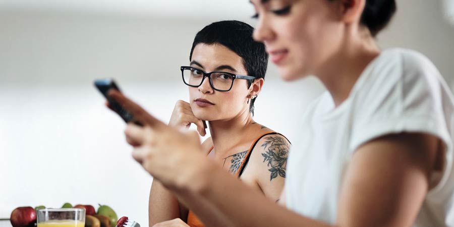 Woman with short cropped hair looking across at her female partner jealously who is on her mobile 