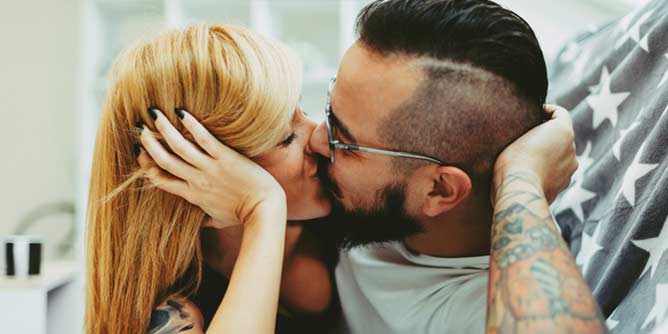 Sexy man with arm tattooes kisses his blonde partner as they lie side by side