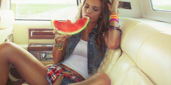 Sexy young woman sitting in the back of a car eating a huge slice of watermelon