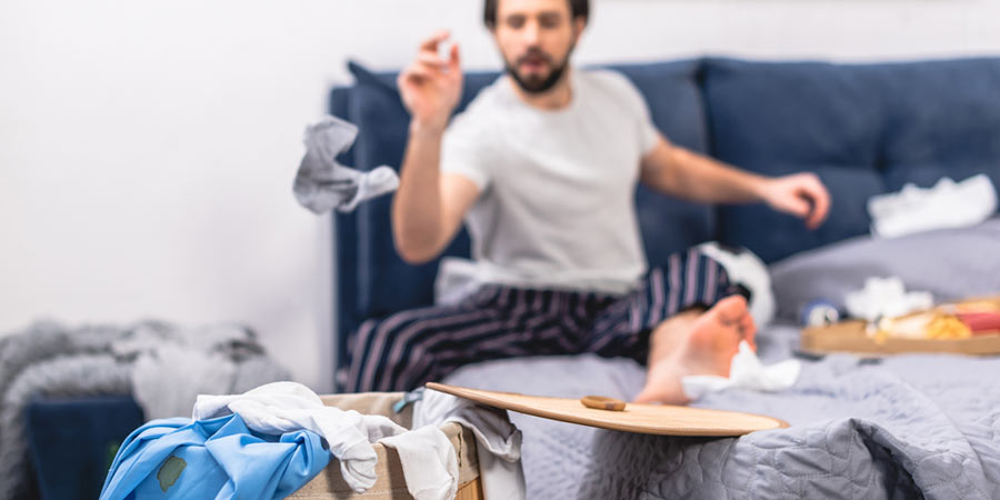 A man in his pyjamas sitting on his bed amongst clutter in a messy bedroom