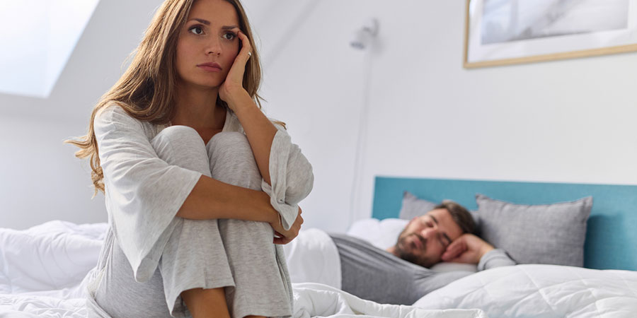 Woman sitting on the edge of a bed looking disappointed whilst her partner is sleeping