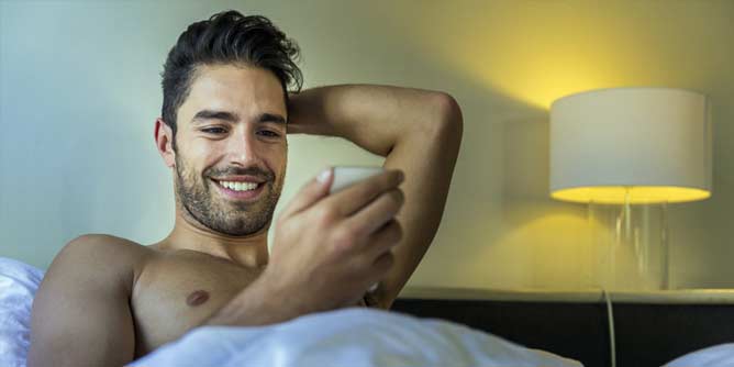 Sexy man with stubble in bed looking at his phone choosing a photo to send his girlfriend