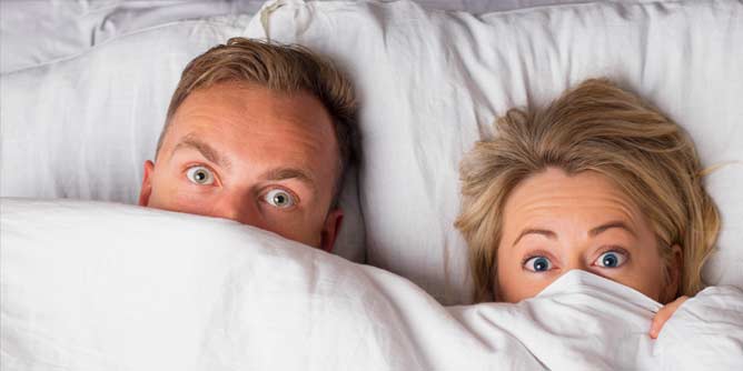 Couple in bed pulling the covers up to hide their faces and looking surprised