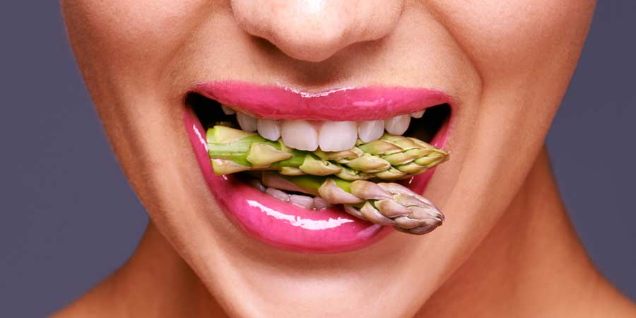Woman's mouth with shiny hot pink lipstick biting into two asparagus spears
