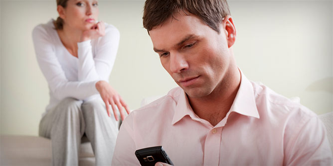 Man using his mobile to cheat whilst his partner is in background looking unhappy