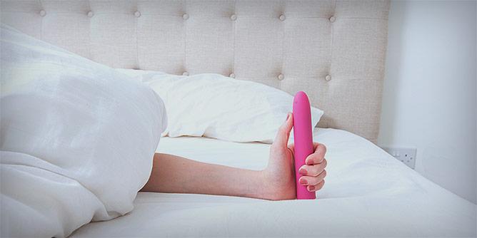 Woman's hand peeking out from under the doona holding a bright pink vibrator