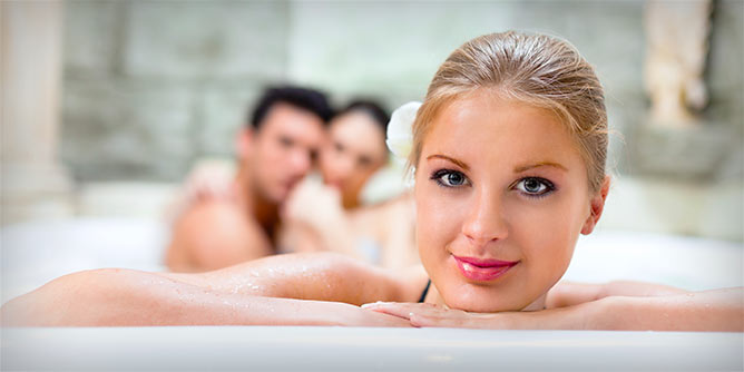 Young blonde woman at side of spa whilst a swinger couple embrace behind her