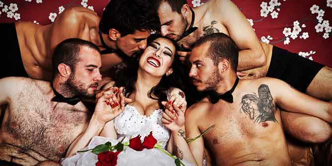 Smiling woman wearing a wedding gown surrounded by bare chested handsome men